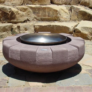 May include: A large, round, stone fire pit with a metal bowl insert. The fire pit is made of a rough, textured stone and has a slightly rounded top. The metal bowl is smooth and shiny.