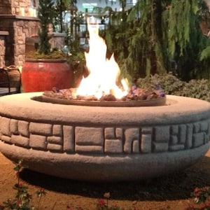 May include: A stone fire pit with a burning flame. The fire pit is circular with a decorative, rectangular pattern around the base. The flames are bright and surrounded by decorative rocks.