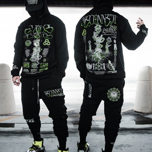 Op de afbeelding: Een zwarte hoodie met een groene en witte grafische print met de tekst "Reconnect with your Source" en "Evolve Your Trinity". De hoodie wordt gecombineerd met een zwarte joggingbroek met een vergelijkbare grafische print op het linkerbeen.