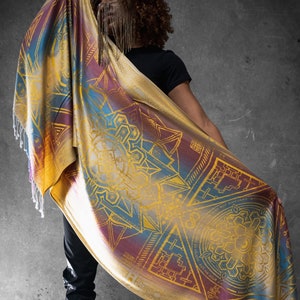 Può includere: Una persona che indossa una camicia nera e tiene un grande scialle colorato con un motivo geometrico in oro. Lo scialle presenta un gradiente di colori, tra cui giallo, arancione, rosso, blu e viola. Il motivo è intricato e include una varietà di forme, tra cui triangoli, quadrati e cerchi.