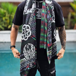 May include: A black scarf featuring white and multi-coloured geometric and abstract patterns. The scarf, with a fringed edge, is draped over a black t-shirt. The design includes chains, floral motifs, and a central graphic.