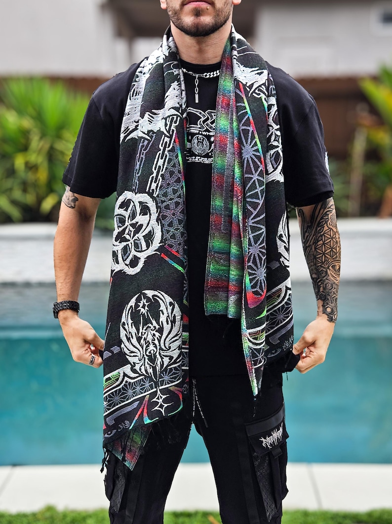 May include: A black scarf featuring white and multi-coloured geometric and abstract patterns. The scarf, with a fringed edge, is draped over a black t-shirt. The design includes chains, floral motifs, and a central graphic.
