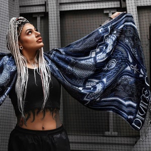 May include: A woman with white braids wears a black crop top and black pants. She is holding a blue and white patterned scarf with a geometric design and the word "Divinity" printed on it.