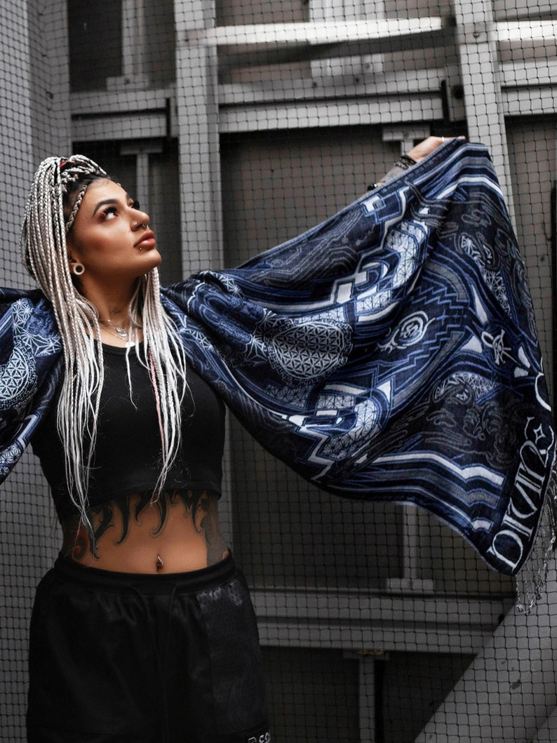 May include: A woman with white braids wears a black crop top and black pants. She is holding a blue and white patterned scarf with a geometric design and the word "Divinity" printed on it.