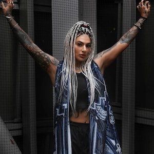 May include: A woman with long white braids wears a black tank top and black pants. She is wearing a blue and white scarf with a graphic print. The scarf has the word "NATION" printed on it.