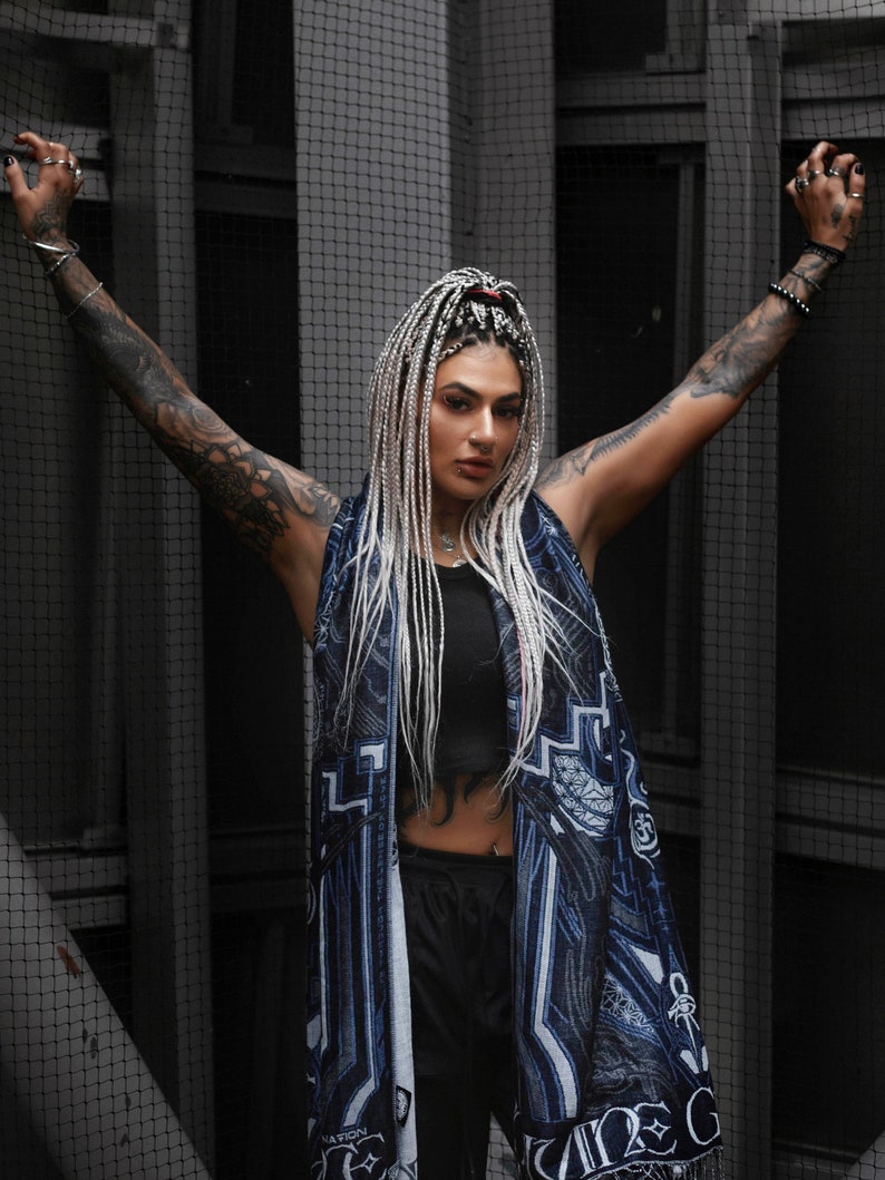 May include: A woman with long white braids wears a black tank top and black pants. She is wearing a blue and white scarf with a graphic print. The scarf has the word "NATION" printed on it.