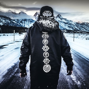 Puede incluir: Una chaqueta negra con capucha con un gráfico blanco de "AWAKEN" en la capucha y una fila vertical de símbolos de chakras blancos en la espalda. La chaqueta la lleva una persona que lleva una gorra de béisbol negra y blanca con el texto "DREAM AWAKZ" en la parte delantera.