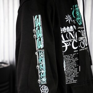 May include: Black hooded sweatshirt with a graphic print on the front and sleeves. The print features a combination of Japanese characters, abstract shapes, and text. The text on the sleeve reads "ASSONNTEN GRONTORET" and the text on the front reads "SCHOLD THE GIFT OF LIFE AND THE CHANCE TO EXPERIENCE THE WORLD THROUGH MANY LIVES CONSTRIUTLY RENJEUTING OURSELVES IN THE PURSUIT OF HAPPINESS AND UNDERSTANDING CXSTETICE HERE MAY NOT BE EASY BUT ITS BEAUTY TRANSCENDS ALL STACLES WELCOME TO THE GREAT AWAKENING".