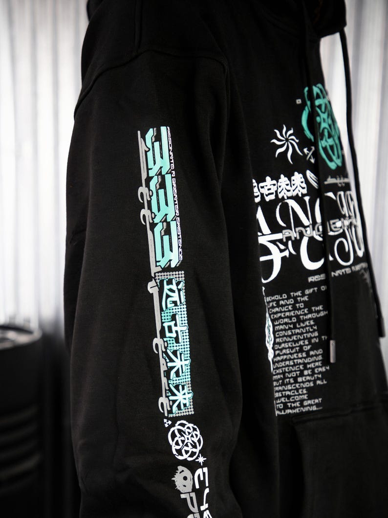May include: Black hooded sweatshirt with a graphic print on the front and sleeves. The print features a combination of Japanese characters, abstract shapes, and text. The text on the sleeve reads "ASSONNTEN GRONTORET" and the text on the front reads "SCHOLD THE GIFT OF LIFE AND THE CHANCE TO EXPERIENCE THE WORLD THROUGH MANY LIVES CONSTRIUTLY RENJEUTING OURSELVES IN THE PURSUIT OF HAPPINESS AND UNDERSTANDING CXSTETICE HERE MAY NOT BE EASY BUT ITS BEAUTY TRANSCENDS ALL STACLES WELCOME TO THE GREAT AWAKENING".