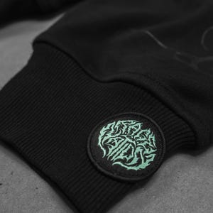 May include: Black sweatshirt with a green embroidered patch on the cuff. The patch features a stylized design with the text "SLAUGHTER"