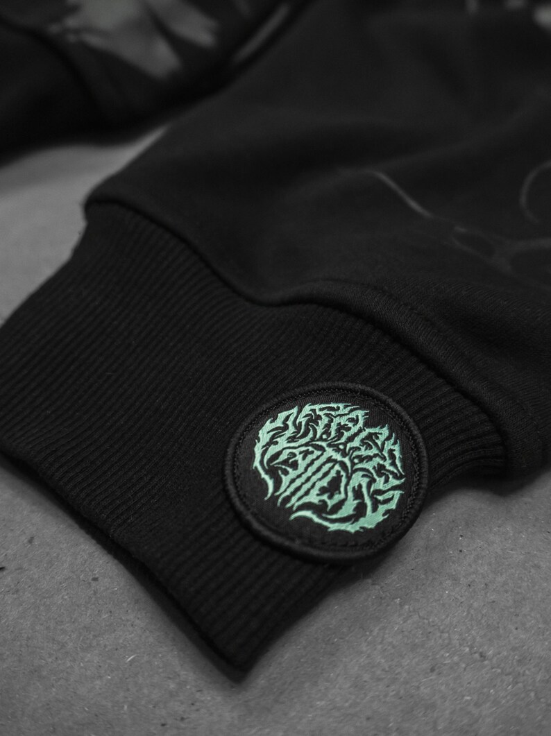 May include: Black sweatshirt with a green embroidered patch on the cuff. The patch features a stylized design with the text "SLAUGHTER"