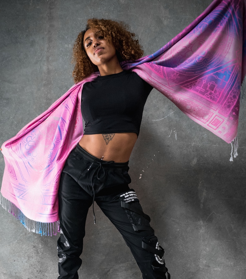 May include: A woman wearing black pants and a black crop top with a pink and purple patterned scarf draped around her shoulders. The scarf has a geometric design and a fringe.