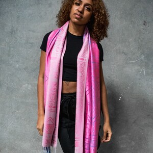 May include: A pink and blue patterned scarf with geometric designs. The scarf is draped around the neck of a person wearing a black top and black pants.