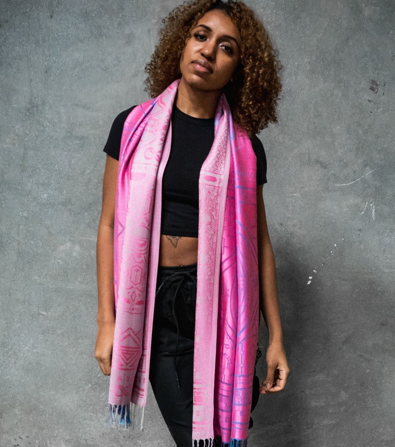 May include: A pink and blue patterned scarf with geometric designs. The scarf is draped around the neck of a person wearing a black top and black pants.