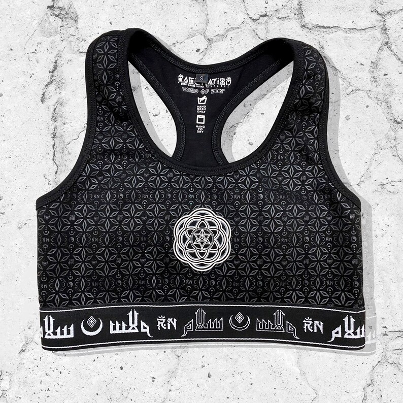 May include: Black sports bra with a geometric pattern and a white mandala design. The bra has a black band with white Arabic script that reads "Salam" on the bottom.
