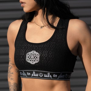 May include: Black sports bra with a white geometric pattern and a white band with Arabic script. The bra has a scoop neck and racerback style.