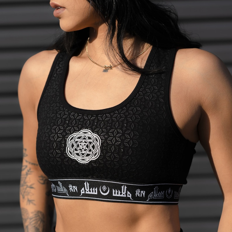 May include: Black sports bra with a white geometric pattern and a white band with Arabic script. The bra has a scoop neck and racerback style.