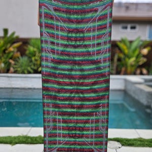 May include: A rectangular textile with a striped pattern in shades of green, red, and grey, with white geometric designs. The textile is held up, showcasing its full length. The edges have a fringe detail.