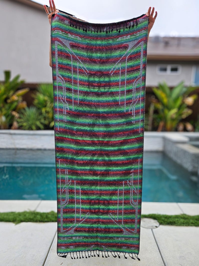 May include: A rectangular textile with a striped pattern in shades of green, red, and grey, with white geometric designs. The textile is held up, showcasing its full length. The edges have a fringe detail.