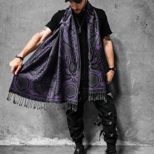 May include: A black and purple scarf with a detailed pattern featuring hands, eyes, and geometric shapes. The scarf has a fringe along the bottom edge. The scarf is draped over a black shirt.