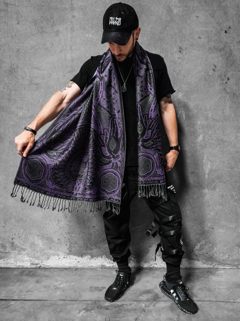 May include: A black and purple scarf with a detailed pattern featuring hands, eyes, and geometric shapes. The scarf has a fringe along the bottom edge. The scarf is draped over a black shirt.
