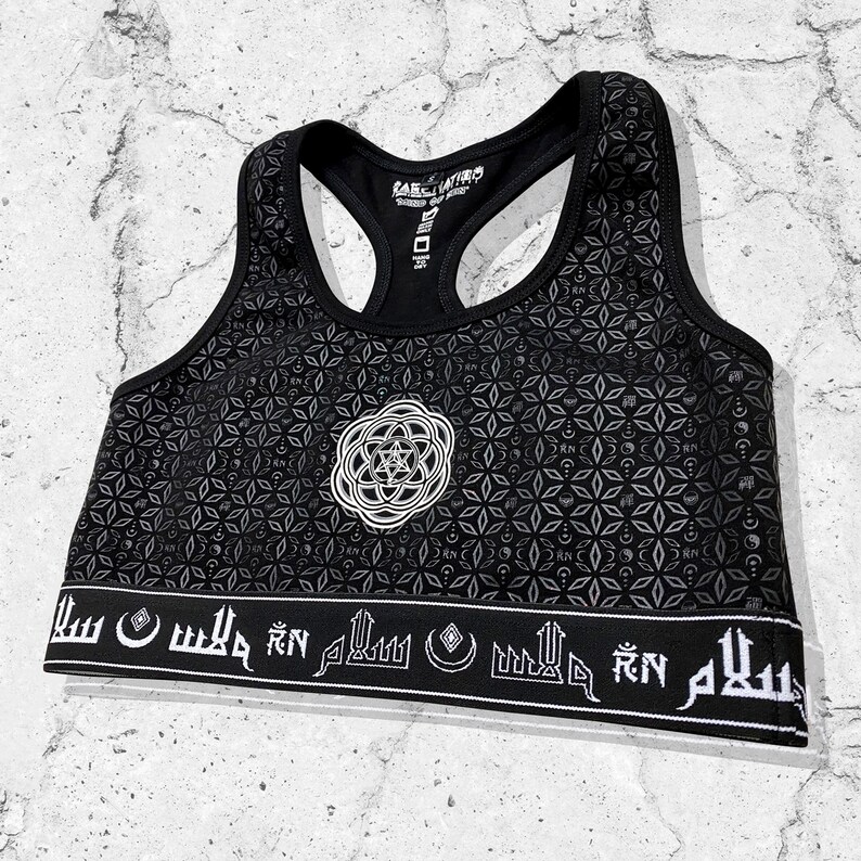May include: Black sports bra with a white geometric pattern and a white circular design in the center. The bottom band has white Arabic script.