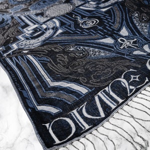 May include: A blue and gray patterned scarf with a fringe. The scarf has the word "DIVINE" woven into the fabric.