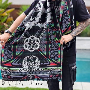 May include: Black scarf with a detailed geometric and fantasy design. The scarf features a repeating pattern of chains, geometric shapes, and a central emblem with wings. The edges are trimmed with a rainbow gradient and tassels.