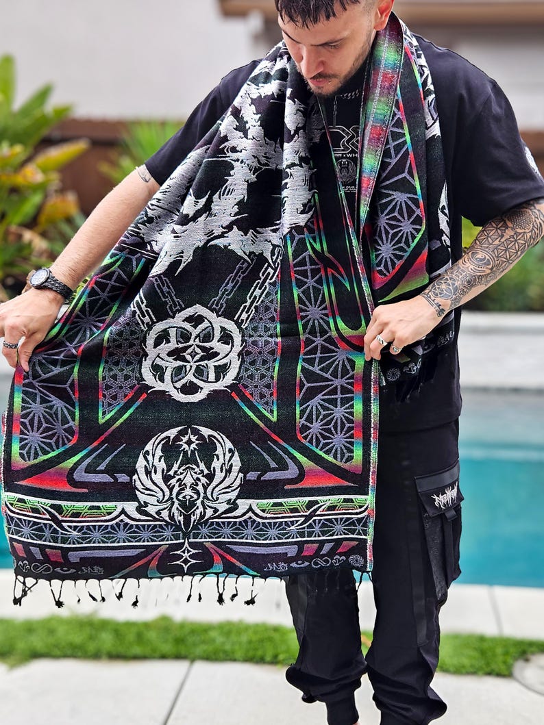 May include: Black scarf with a detailed geometric and fantasy design. The scarf features a repeating pattern of chains, geometric shapes, and a central emblem with wings. The edges are trimmed with a rainbow gradient and tassels.