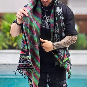 May include: A patterned scarf in shades of red, green, and black is worn over a black top. The scarf features a geometric design and fringed edges. The person is also wearing black trousers. The scarf's design incorporates various patterns and colours.