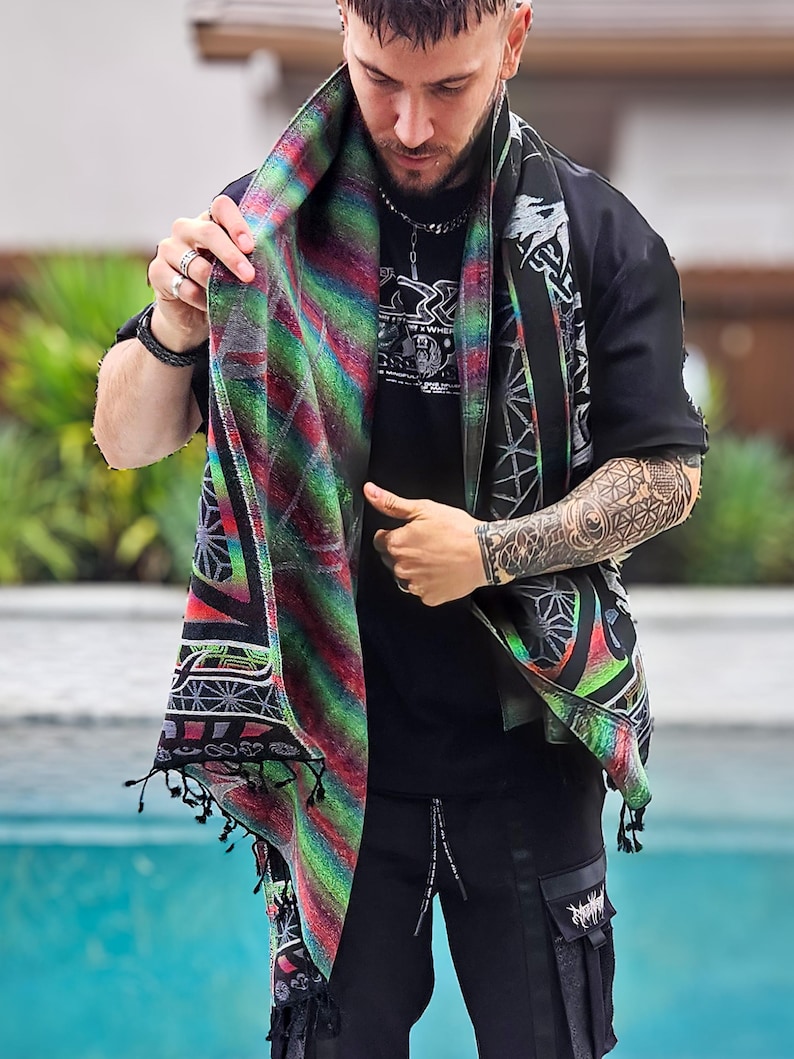 May include: A patterned scarf in shades of red, green, and black is worn over a black top. The scarf features a geometric design and fringed edges. The person is also wearing black trousers. The scarf's design incorporates various patterns and colours.