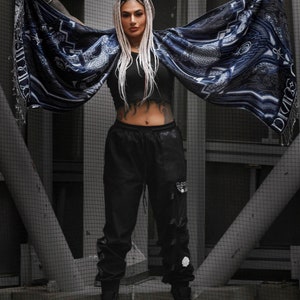 May include: A person wearing black pants and a blue and white patterned shawl with a geometric design. The person has long white hair and is standing in front of a metal grate.