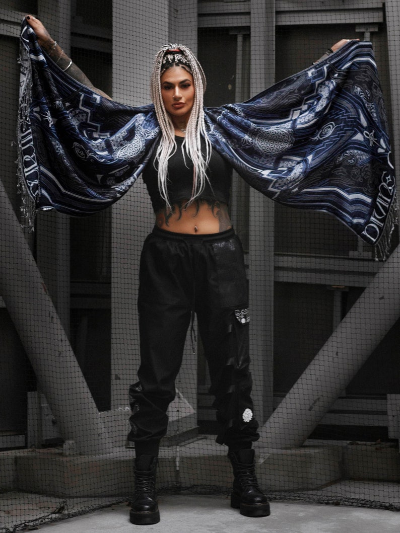 May include: A person wearing black pants and a blue and white patterned shawl with a geometric design. The person has long white hair and is standing in front of a metal grate.