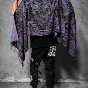 May include: A black and purple shawl featuring geometric and eye patterns. The shawl has a fringed edge. The person in the image is wearing black trousers with multiple straps and buckles.