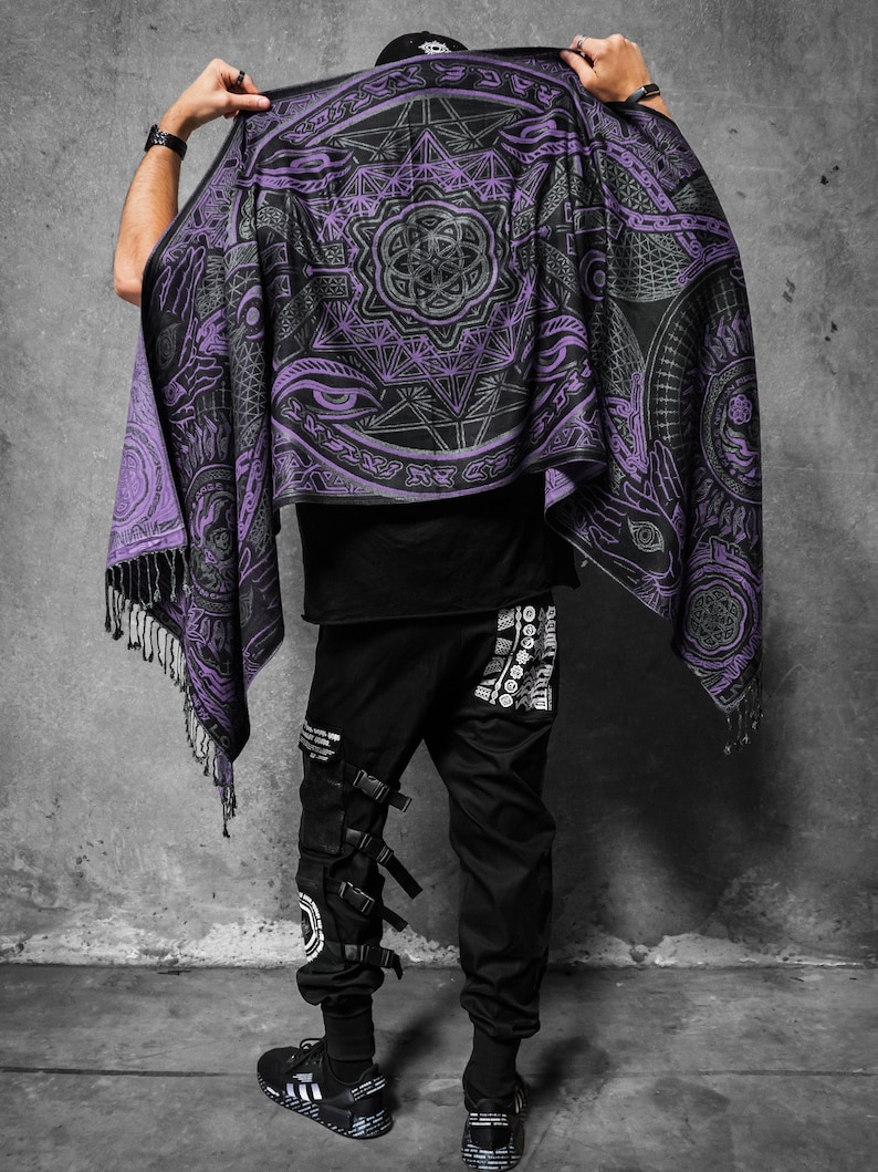 May include: A black and purple shawl featuring geometric and eye patterns. The shawl has a fringed edge. The person in the image is wearing black trousers with multiple straps and buckles.