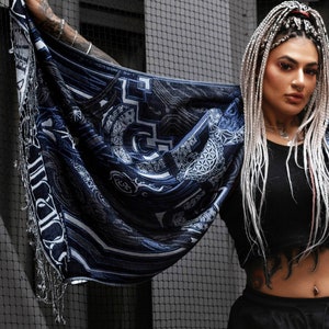 May include: A woman with long white braids wears a black top and black pants. She is holding a blue and white patterned scarf with intricate designs. The scarf has a fringe border.