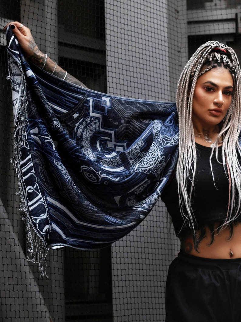 May include: A woman with long white braids wears a black top and black pants. She is holding a blue and white patterned scarf with intricate designs. The scarf has a fringe border.