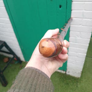 Puede incluir: Un bastón de madera con un mango redondeado y un acabado marrón oscuro. El bastón tiene una serie de pequeños acentos oscuros a lo largo de su longitud. El mango es de un tono marrón más claro con un patrón de vetas de madera visible.