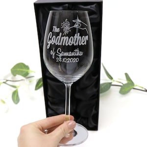 May include: A clear glass wine glass with an engraved design. The design includes a hand holding a wand with stars coming out of it. The text reads "The Godmother of Samantha 24.10.2020".
