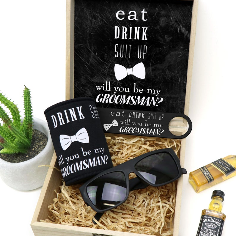 May include: A wooden box filled with a black can cooler, sunglasses, a bottle opener, and a miniature bottle of liquor. The can cooler, bottle opener, and sunglasses all have the text "eat drink suit up will you be my groomsman?" printed on them.