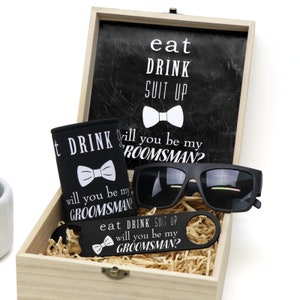 May include: A wooden box with a black and white sign that says "eat drink suit up" and a bow tie. The box contains a black can cooler with a bow tie and the text "will you be my groomsman?" A pair of black sunglasses, a black bottle opener with the text "eat drink suit up will you be my groomsman?" and a bow tie.