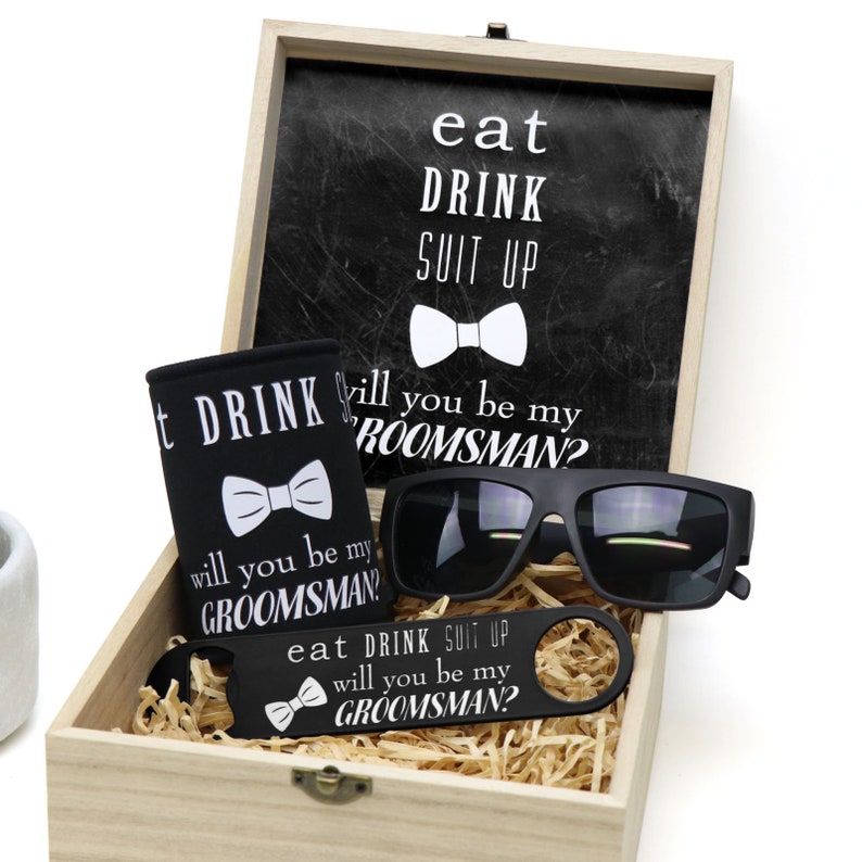 May include: A wooden box with a black and white sign that says "eat drink suit up" and a bow tie. The box contains a black can cooler with a bow tie and the text "will you be my groomsman?" A pair of black sunglasses, a black bottle opener with the text "eat drink suit up will you be my groomsman?" and a bow tie.