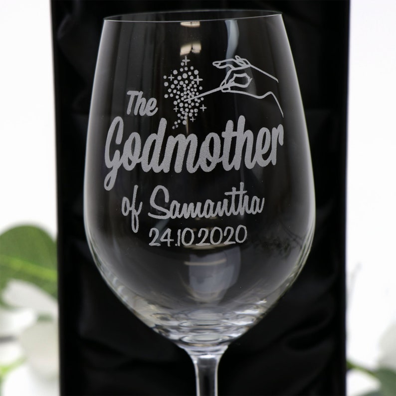 May include: A clear glass wine glass with an etched design that says "The Godmother of Samantha 24.10.2020". The design includes a hand holding a wand with sparkles.