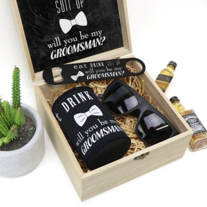 May include: A wooden box with a black lid that says "Suit Up, Will You Be My Groomsman?" The box contains a black can cooler with a bow tie and the text "Eat Drink Suit Up, Will You Be My Groomsman?" A black bottle opener with the same text, a pair of black sunglasses, and two miniature bottles of liquor.