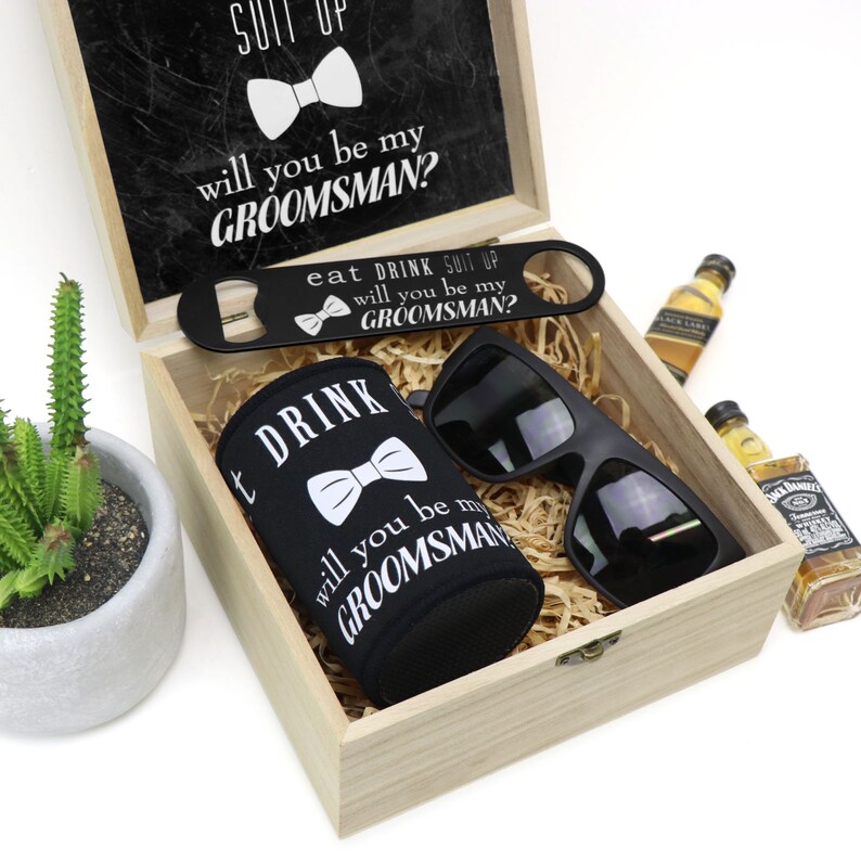 May include: A wooden box with a black lid that says "Suit Up, Will You Be My Groomsman?" The box contains a black can cooler with a bow tie and the text "Eat Drink Suit Up, Will You Be My Groomsman?" A black bottle opener with the same text, a pair of black sunglasses, and two miniature bottles of liquor.