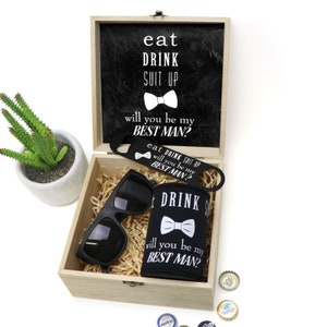 May include: A wooden box with a black chalkboard lid that says "eat drink suit up will you be my best man?" The box contains a black can cooler with a bow tie and the text "eat drink suit up will you be my best man?" a pair of black sunglasses, and a bottle opener with the same text.