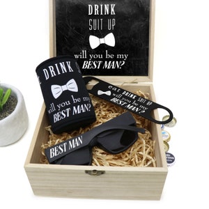 May include: A wooden box with a black chalkboard lid that says "Drink Suit Up, Will You Be My Best Man?" The box contains a black can cooler with the same text, a black bottle opener with the text "Eat Drink Suit Up, Will You Be My Best Man?", a pair of black sunglasses with the text "Best Man" on the lenses, and a brown paper filler.