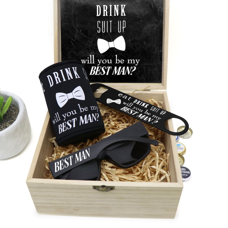 May include: A wooden box with a black chalkboard lid that says "Drink Suit Up, Will You Be My Best Man?" The box contains a black can cooler with the same text, a black bottle opener with the text "Eat Drink Suit Up, Will You Be My Best Man?", a pair of black sunglasses with the text "Best Man" on the lenses, and a brown paper filler.