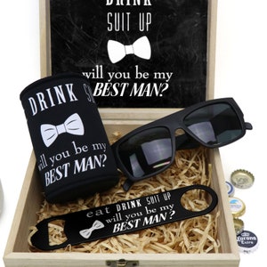 May include: A wooden box with a black chalkboard lid that says "Drink Suit Up, Will You Be My Best Man?" The box contains a black can cooler with the same text, a pair of black sunglasses, a black bottle opener with the same text, and a few bottle caps.
