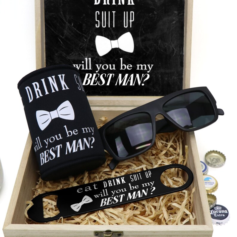 May include: A wooden box with a black chalkboard lid that says "Drink Suit Up, Will You Be My Best Man?" The box contains a black can cooler with the same text, a pair of black sunglasses, a black bottle opener with the same text, and a few bottle caps.